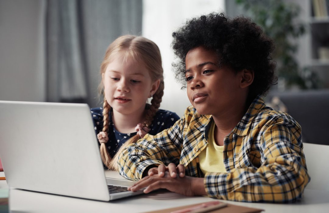 Two children sitting at table in front of laptop looking at monitor during online video call