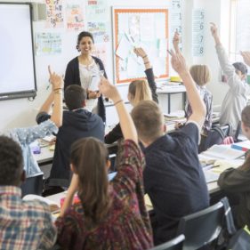 Rear view of teenage students raising hands in classroom